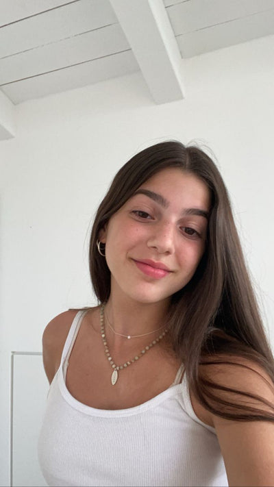 Woman wearing a white tank top and gold necklace against a white wall.