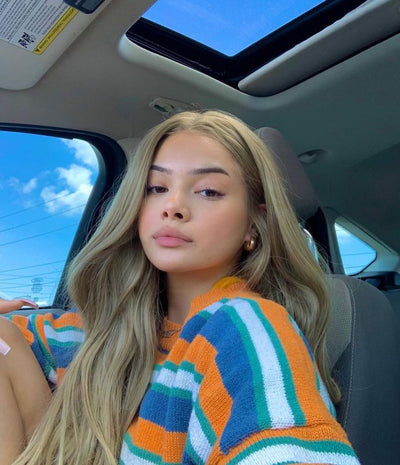 Person sitting in a car wearing a colorful striped sweater with a blue sky visible through the window.